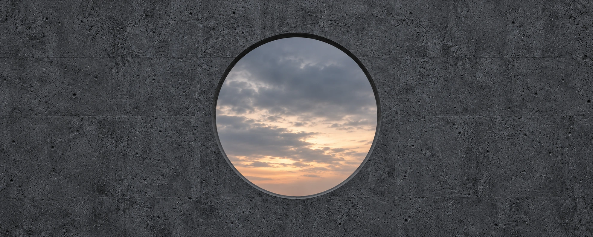 Abertura circular centrada en un muro de concreto gris oscuro, con un cielo suave al atardecer.